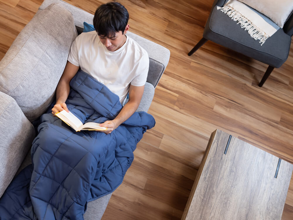 Man reading a book on the couch, with Classic Weighted Blanket draped over his lower body Man reading a book on the couch, with Classic Weighted Blanket draped over his lower body