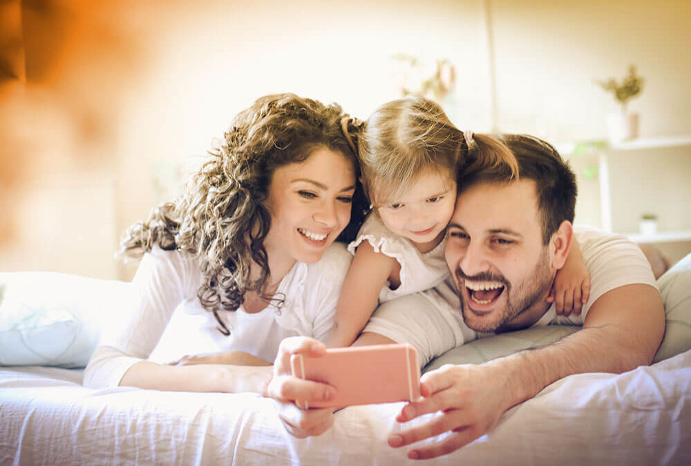 Husband, wife, and young daughter all lying in bed and smiling at something they are seeing on a smartphone