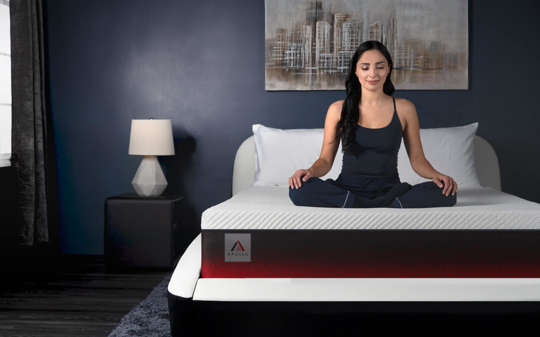 Woman in a bedroom wearing blue pyjamas. She is sitting cross-legged in a meditation pose on top of the Apollo mattress