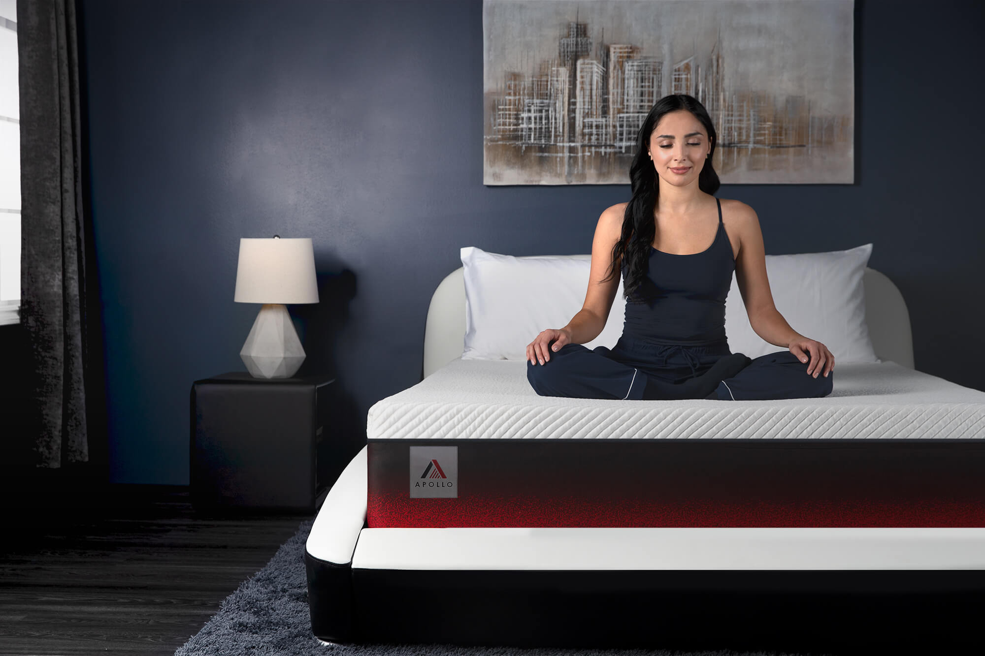 Woman in a bedroom wearing blue pyjamas. She is sitting cross-legged in a meditation pose on top of the Apollo mattress