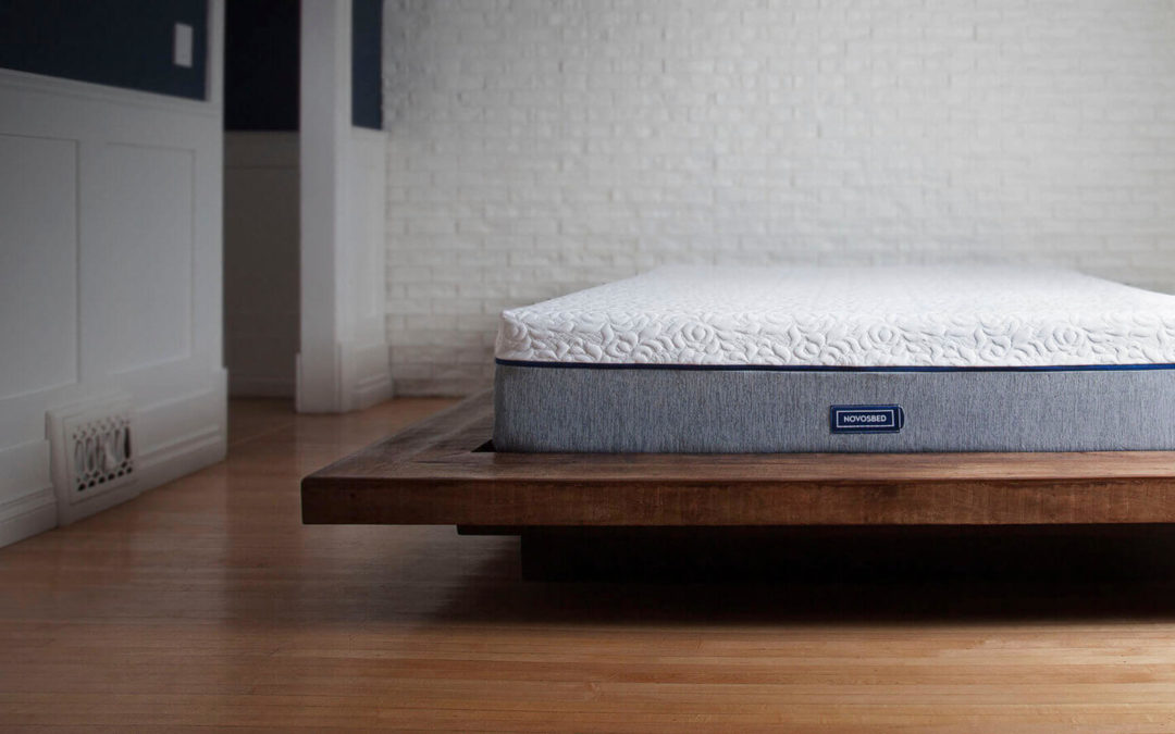 Novosbed premium memory foam mattress resting on a brown platform bed frame in a stylish white brick bedroom