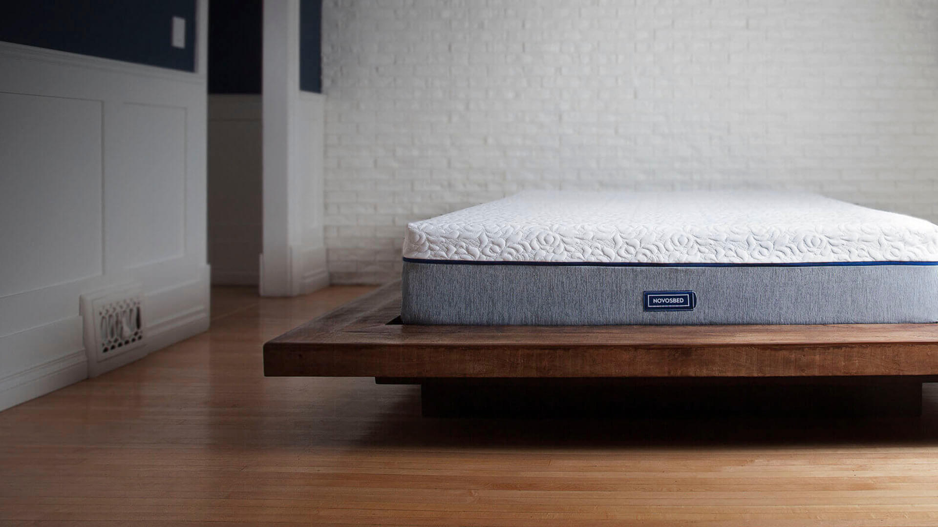 Novosbed premium memory foam mattress resting on a brown platform bed frame in a stylish white brick bedroom