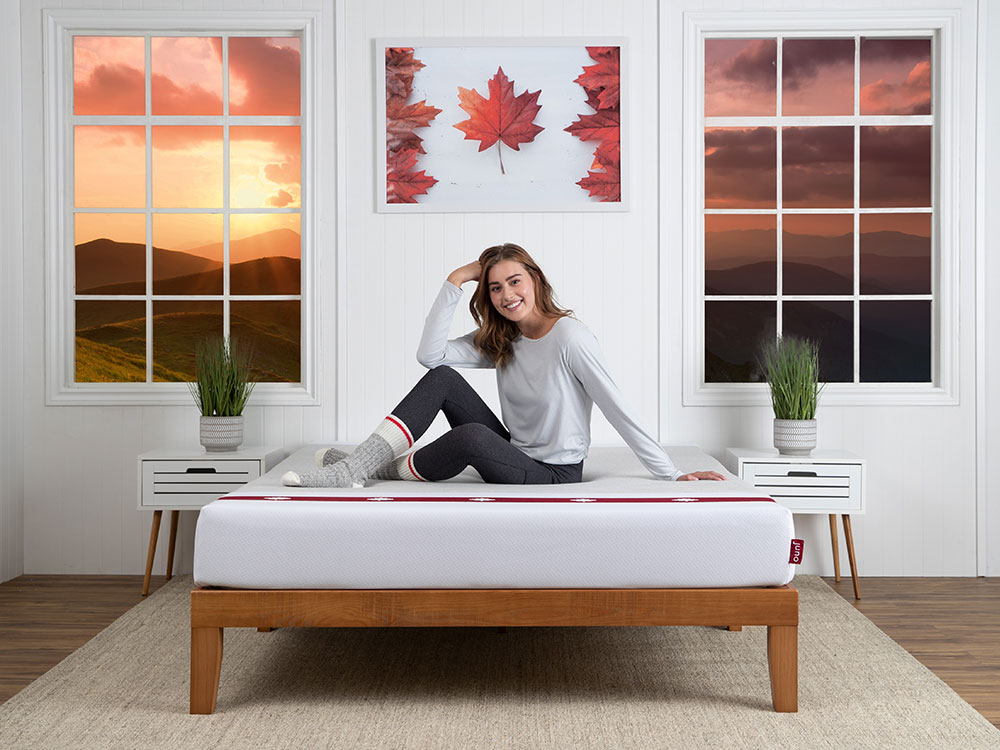 Woman in her bedroom, sitting on the Juno mattress and smiling at the camera. There is an image of the Canadian flag framed and hanging above her bed.