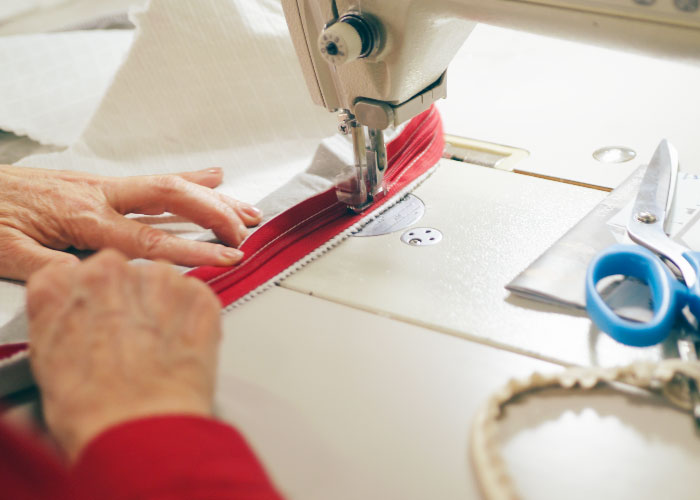 Image of a factory seamstress in a red shirt carefully using a sewing machine to attach the red zipper to the white cover of the Douglas mattress, with blue scissors and other tools within reach.