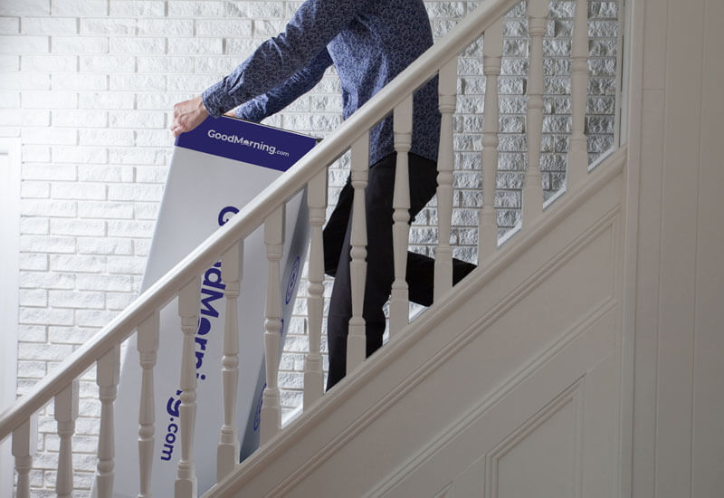 Man pulling a GoodMorning.com mattress box up a white staircase in front of a white brick wall